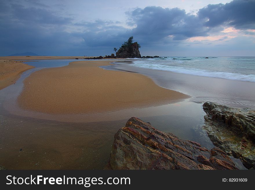 Sea Wave Crashing on Seashore With View of Rock Formation With Trees on Top of It Under Grey Clouds and White Sky