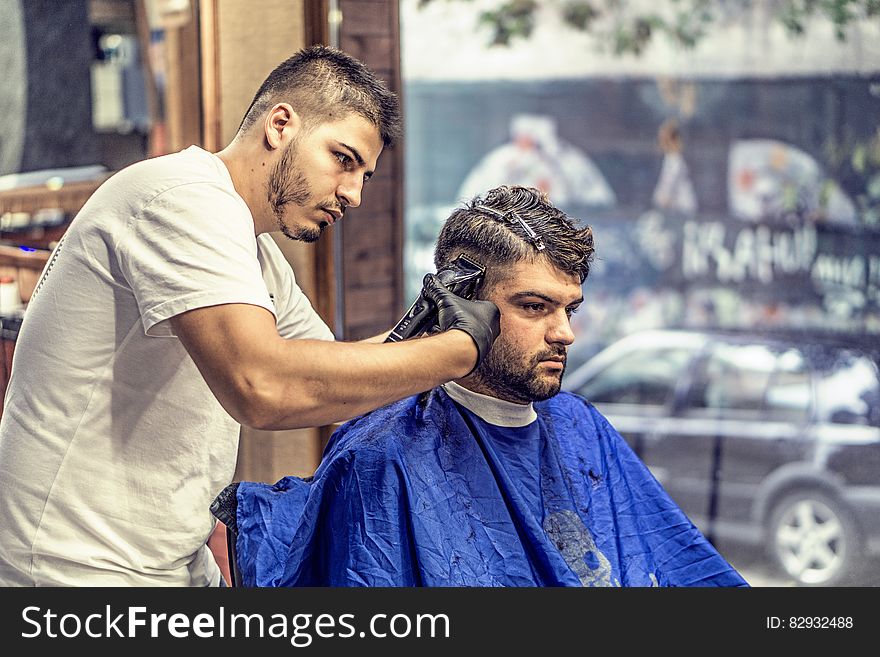Barber in White Shirt Trimming Man&#x27;s Hair in Blue Textile While Sitting Nearby Glass Window