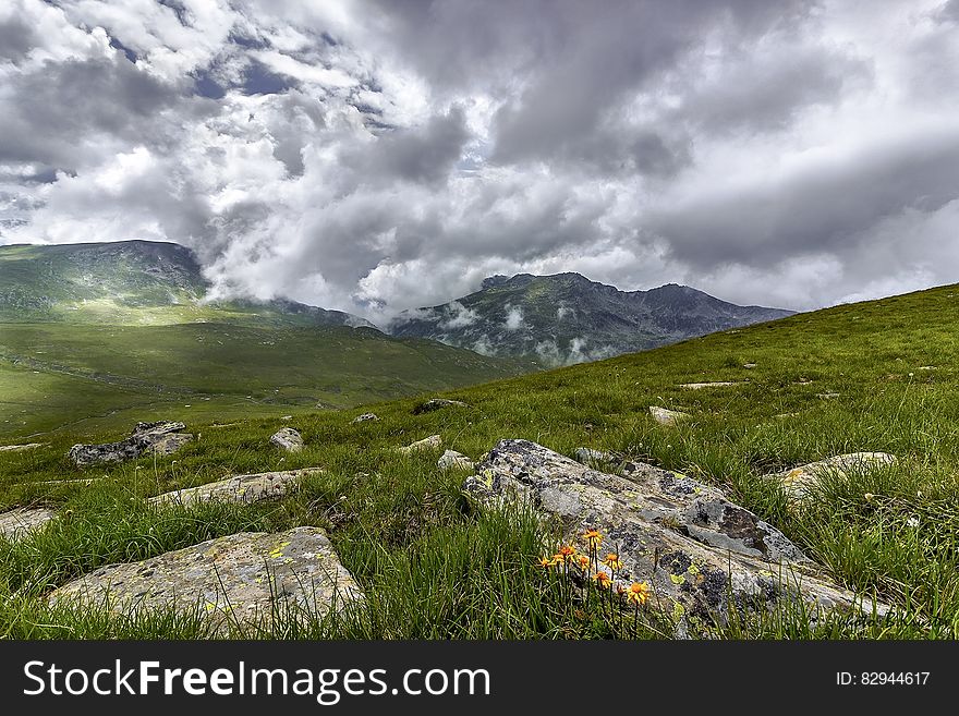 Green Grass Field With Rocks Near Mountains during Cloudy Daytime Sky