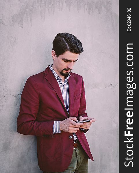 Man Wearing Maroon Blazer Leaning on Gray Concrete Wall While Using His Smartphone