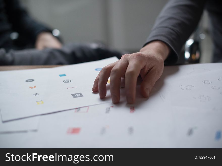 Designer with a hand on sheets of free sample icons from which he must choose (select), office background. Designer with a hand on sheets of free sample icons from which he must choose (select), office background.