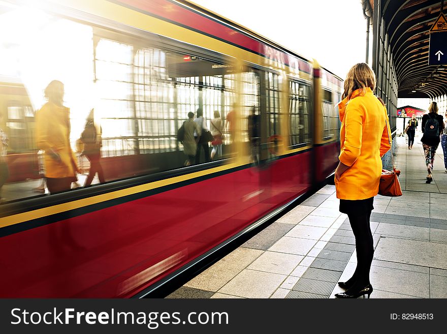 Blond woman in yellow jacket, black skirt and stockings awaits a commuter train in the railway station with one red and yellow train passing with reflections of travelers in the carriage windows. Blond woman in yellow jacket, black skirt and stockings awaits a commuter train in the railway station with one red and yellow train passing with reflections of travelers in the carriage windows.