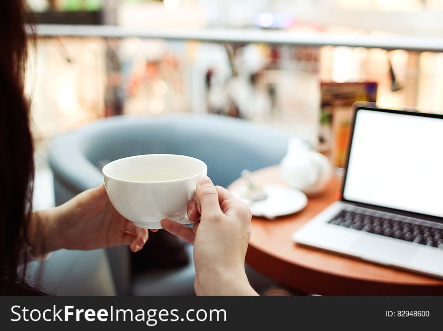 Person Holding White Ceramic Teacup in Front of a Macbook Pro
