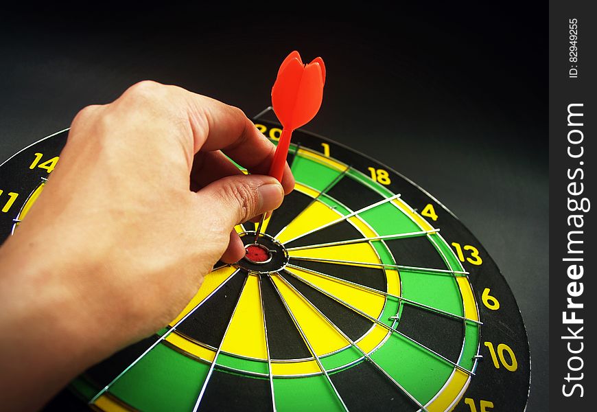 Person Holding Red Dart on Green Yellow and Black Dart Board