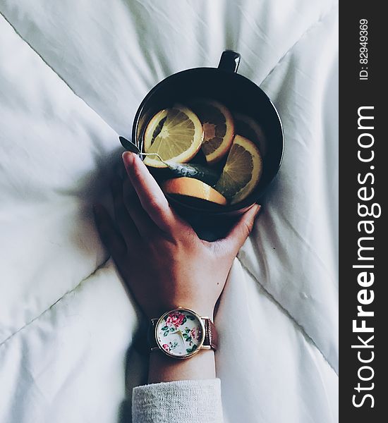 Woman Holding Black Ceramic Mug With Sliced Lemon On Top Of White Bed Comforter
