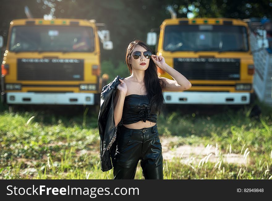 Glamor portrait of dark haired woman posing in black pants and top holding jacket wearing sunglasses outdoor with yellow trucks in field. Glamor portrait of dark haired woman posing in black pants and top holding jacket wearing sunglasses outdoor with yellow trucks in field.
