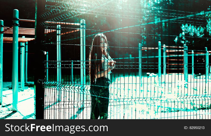 Fashionable young girl stood behind a fence in outdoors playground with bright turquoise lighting highlights.