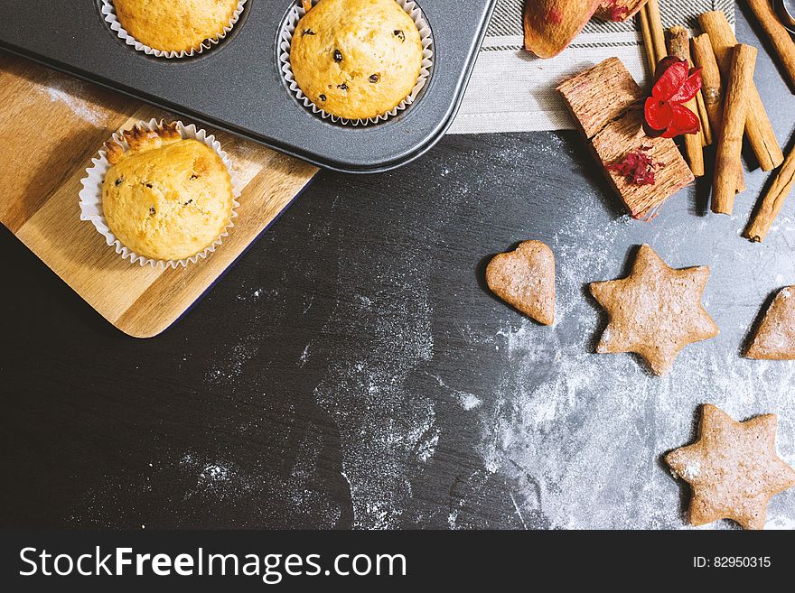 Surface with flour and star shaped biscuits and next to that cup cakes with currents. Other items include cinnamon sticks and a red decorative butterfly are placed alongside. Surface with flour and star shaped biscuits and next to that cup cakes with currents. Other items include cinnamon sticks and a red decorative butterfly are placed alongside.