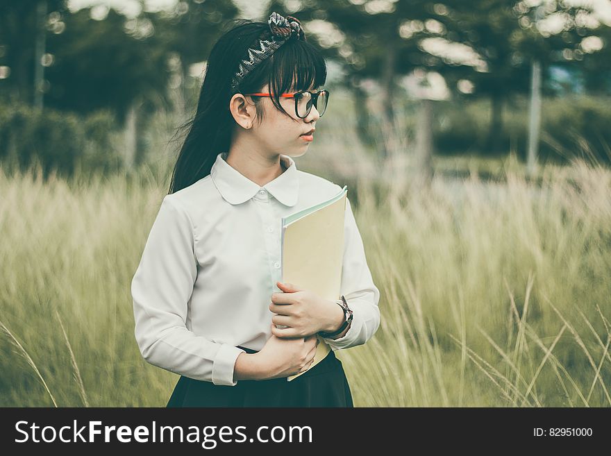 Young attractive Asian schoolgirl with white blouse, dark skirt and spectacles holding school folder while standing outdoors in a hay field, background of dark green trees. Young attractive Asian schoolgirl with white blouse, dark skirt and spectacles holding school folder while standing outdoors in a hay field, background of dark green trees.