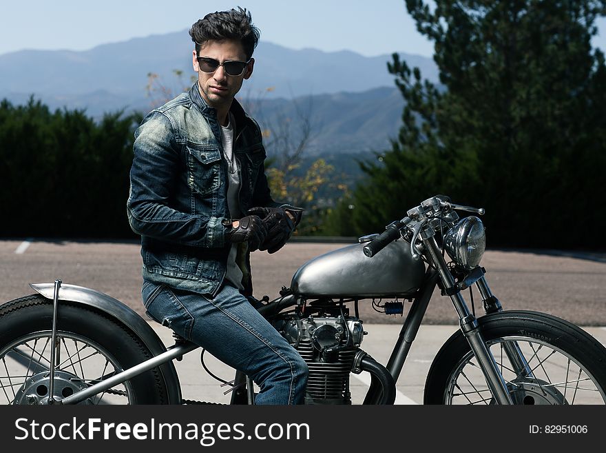 Young man in leather jacket and blue jeans sitting astride a stationary vintage style motorbike with spoke wheels, landscape of dense forest and distant blue mountains. Young man in leather jacket and blue jeans sitting astride a stationary vintage style motorbike with spoke wheels, landscape of dense forest and distant blue mountains.