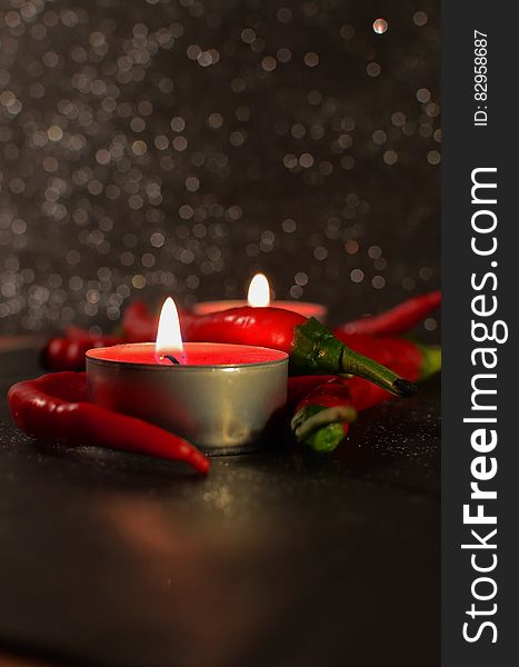 Two burning candles surrounded by red chili peppers on a table. Two burning candles surrounded by red chili peppers on a table.