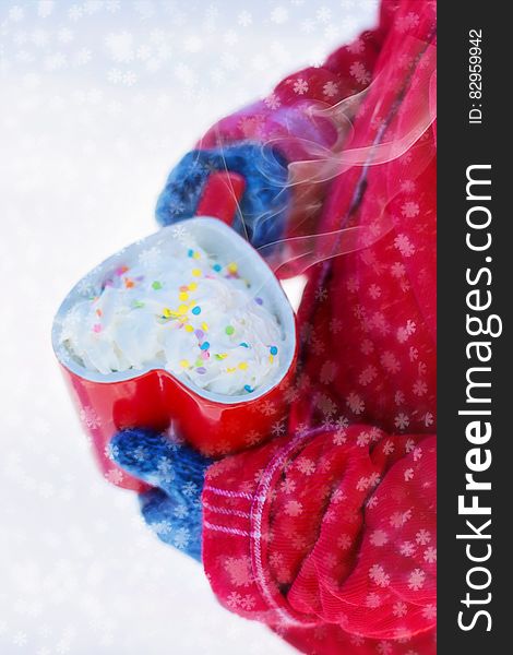 Person in red jacket and blue gloves holding red heart shaped mug of cocoa with whipped cream and sprinkles with snowflakes. Person in red jacket and blue gloves holding red heart shaped mug of cocoa with whipped cream and sprinkles with snowflakes.