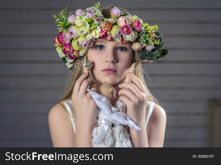 Portrait of young pretty girl wearing floral head-dress composed of many different flowers, white dress with bow and looking pensive, gray slats for a background. Portrait of young pretty girl wearing floral head-dress composed of many different flowers, white dress with bow and looking pensive, gray slats for a background.