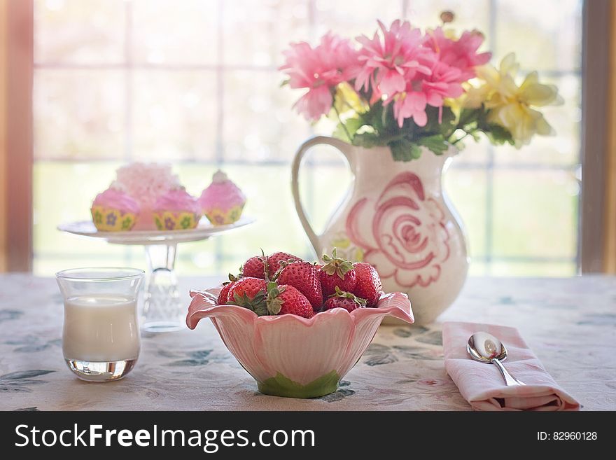 Still life on tabletop with strawberries in pink bowl, glass of milk or cream, plate of cupcakes and vase of flowers against sunny window. Still life on tabletop with strawberries in pink bowl, glass of milk or cream, plate of cupcakes and vase of flowers against sunny window.