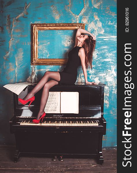 A female model wearing red high heeled shoes sitting on a piano in front of a blue wall. A female model wearing red high heeled shoes sitting on a piano in front of a blue wall.