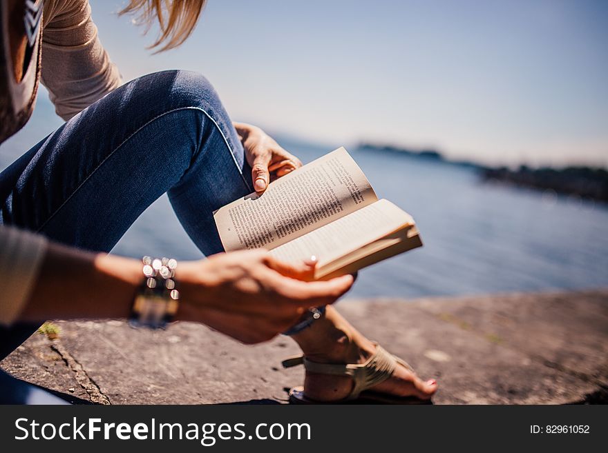 Woman Wearing Blue Denim Jeans Holding Book Sitting on Gray Concrete at Daytime
