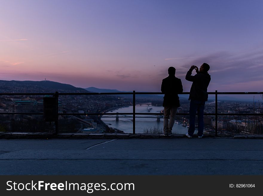 A couple of men standing by a fence looking down into a city. A couple of men standing by a fence looking down into a city.
