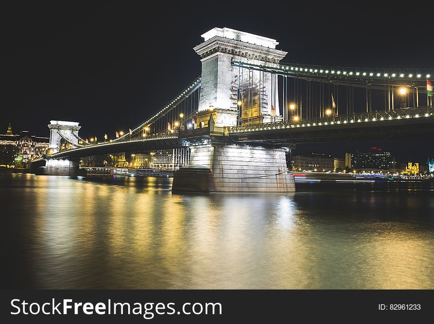 Stone stanchions of suspension bridge illuminated at night reflecting in river along urban waterfront. Stone stanchions of suspension bridge illuminated at night reflecting in river along urban waterfront.