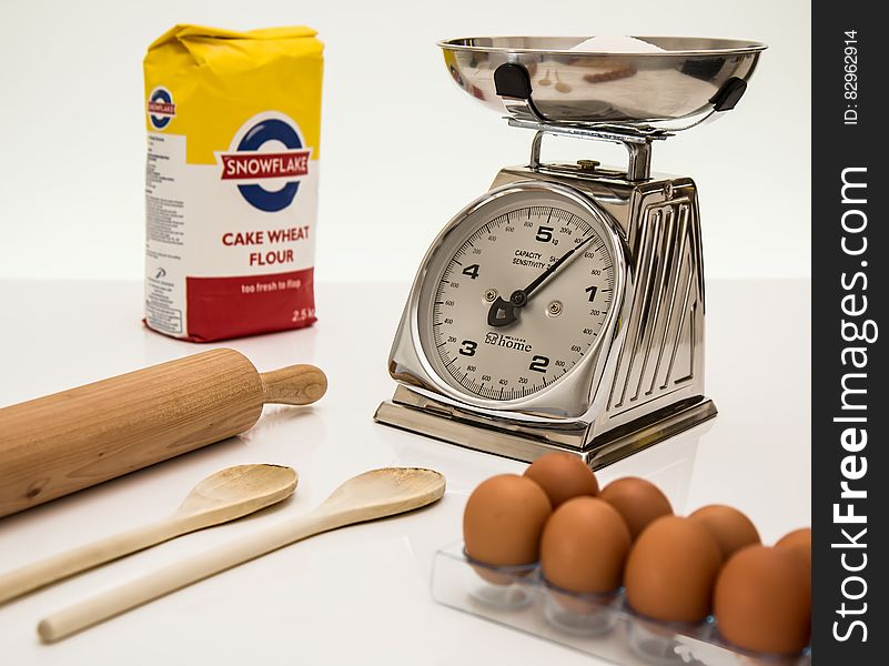 Weighing scales with flour in pan, tray of fresh brown eggs, packet of cake wheat flour, wooden spoons and rolling pin, white background. Weighing scales with flour in pan, tray of fresh brown eggs, packet of cake wheat flour, wooden spoons and rolling pin, white background.