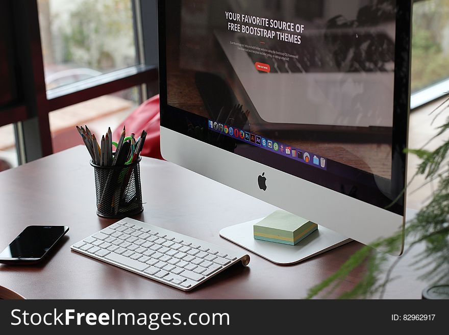 Large screen display and keyboard of Apple computer with memo pad, beaker filled with pencils and pens, office background. Large screen display and keyboard of Apple computer with memo pad, beaker filled with pencils and pens, office background.