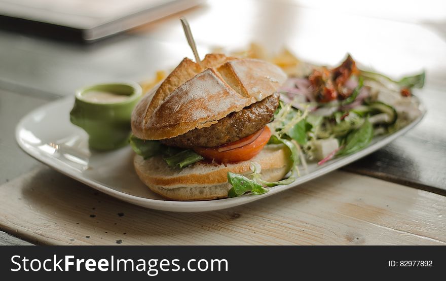 Hamburger on bun with salad on white china plate on tabletop. Hamburger on bun with salad on white china plate on tabletop.