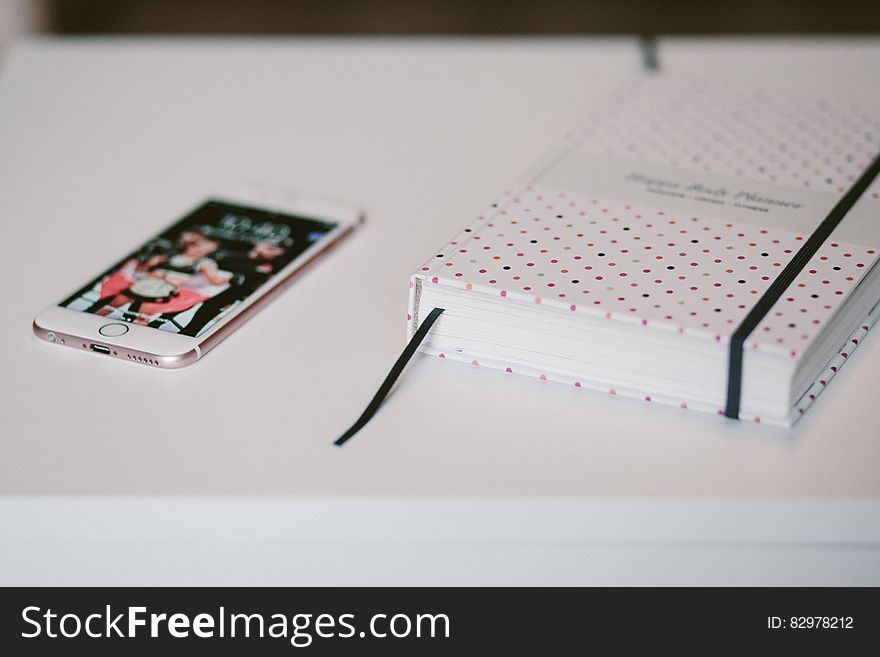 Rose Gold Iphone 6s Beside White And Black Polka Dots Book Both On Top Of White Wooden Table