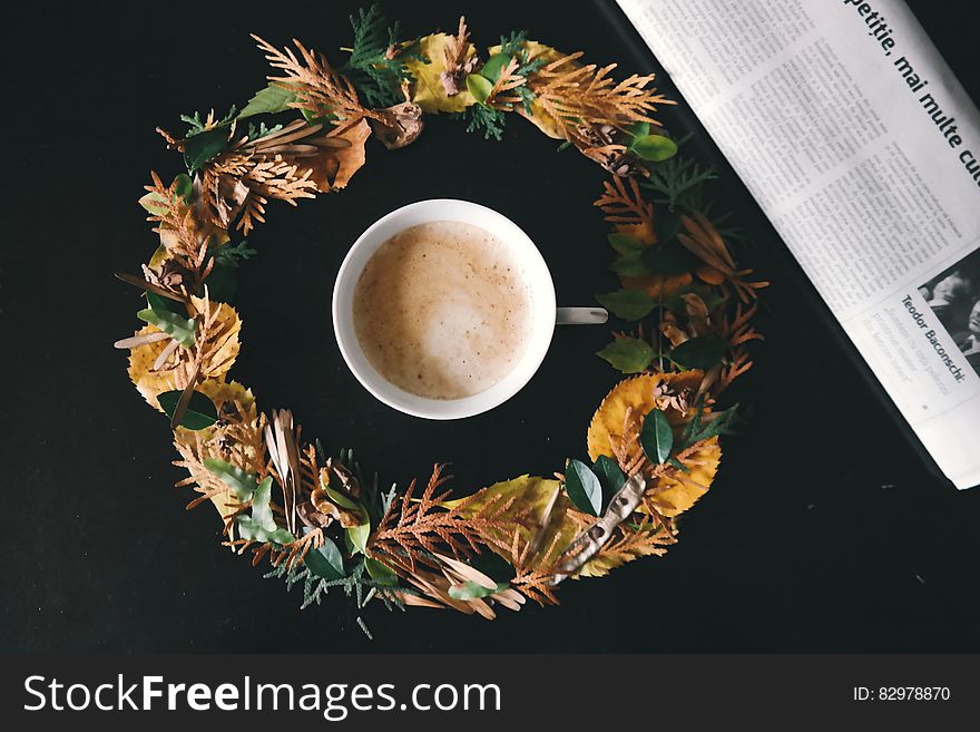A cup of coffee in a wreath of leaves next to a newspaper. A cup of coffee in a wreath of leaves next to a newspaper.