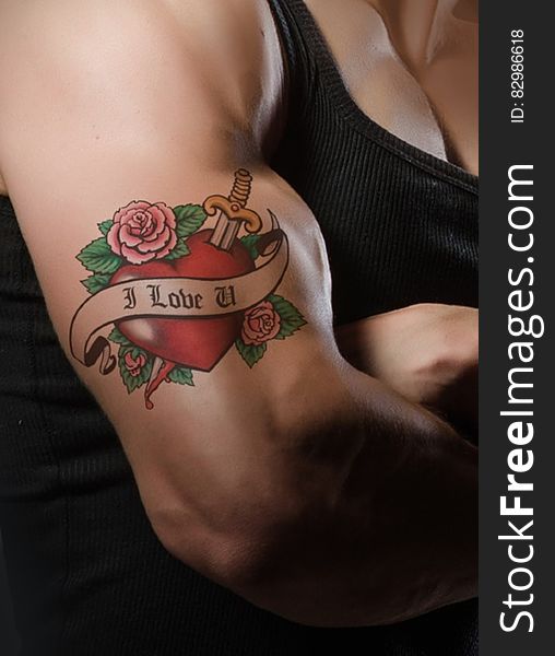 Tattoo on man's muscular arm with a picture of a red heart shape with roses and a dagger embellished with scroll and text "I love you", black background. Tattoo on man's muscular arm with a picture of a red heart shape with roses and a dagger embellished with scroll and text "I love you", black background.