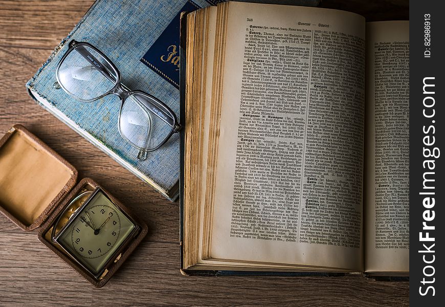 Eyeglasses next to an open old book and a small square clock beside. Eyeglasses next to an open old book and a small square clock beside.