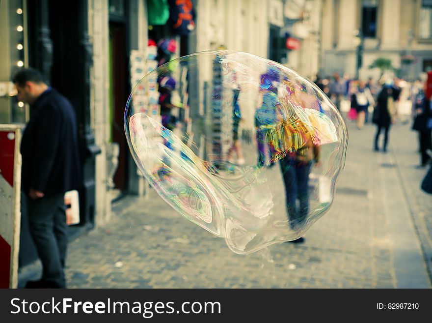 Closeup of very large soap bubble floating in the city street just above the pavement blurring an approaching shopper carrying a carrier bag. Closeup of very large soap bubble floating in the city street just above the pavement blurring an approaching shopper carrying a carrier bag.