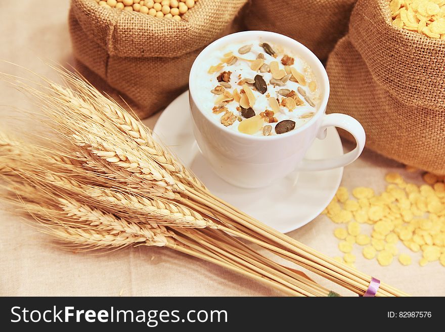 White china cup of coffee decorated on top with nuts and seeds. Placed along side are spikes of corn and behind the coffee are sacks of cereal. White china cup of coffee decorated on top with nuts and seeds. Placed along side are spikes of corn and behind the coffee are sacks of cereal.