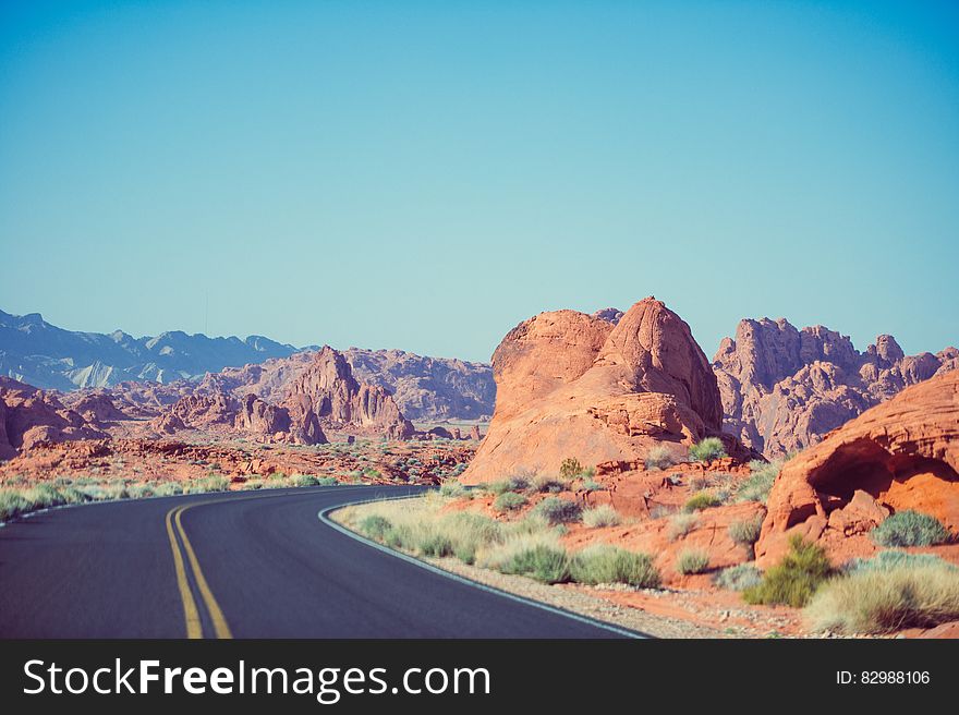 Sharp corner of road with central yellow marking leading through red rocky desert countryside, pale blue sky background. . Sharp corner of road with central yellow marking leading through red rocky desert countryside, pale blue sky background. .
