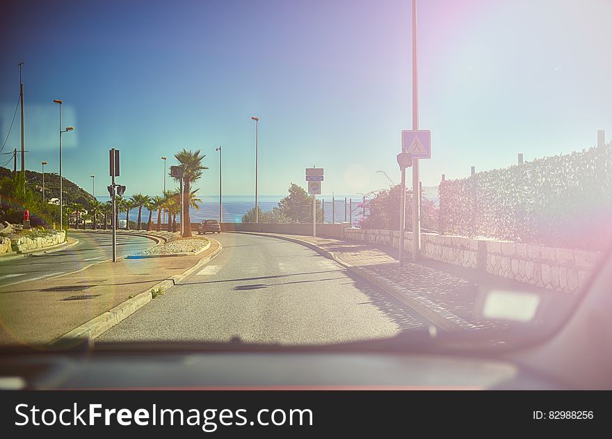 This picture is taken from inside a car through the windhield with the dashboard visible on the lower edge of the picture. The car is driving down a palm-lined ocean road under a blue sky with the ocean inthe background. The sunshine is reflected in the windshield of the car. This picture is taken from inside a car through the windhield with the dashboard visible on the lower edge of the picture. The car is driving down a palm-lined ocean road under a blue sky with the ocean inthe background. The sunshine is reflected in the windshield of the car.