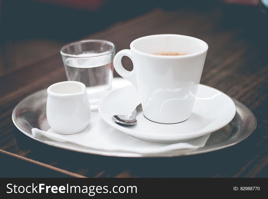 Coffee in a white China cup, cream in a jug, and a glass of water all on a silver tray, placed upon dark tray with wood grain. Coffee in a white China cup, cream in a jug, and a glass of water all on a silver tray, placed upon dark tray with wood grain.