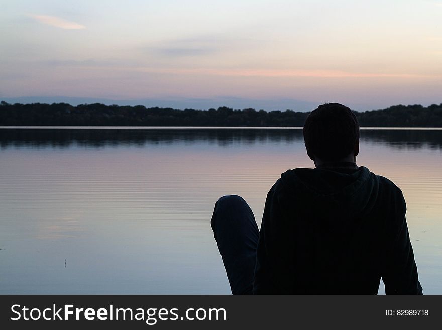 Silhouette of young man sitting on the ground beside a placid lake with forest and pale gray sky in the background. Silhouette of young man sitting on the ground beside a placid lake with forest and pale gray sky in the background.