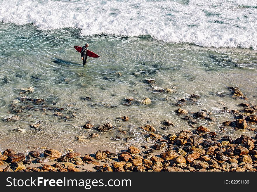 Person Holding Red Surfing Board in Clear Water Near Brown Stone during Daytime