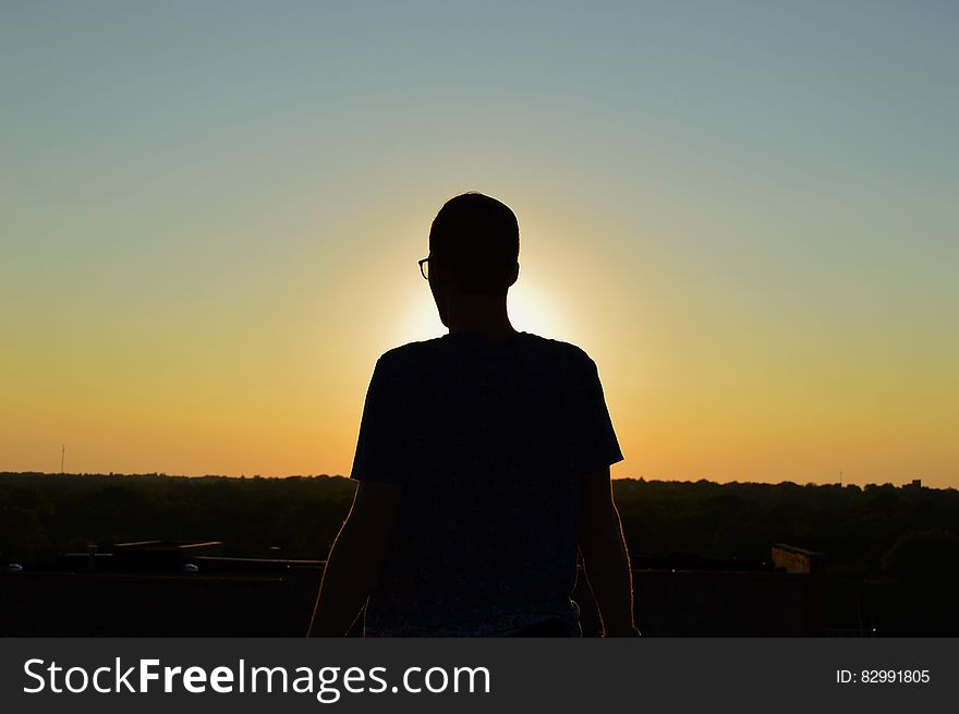 Silhouette of young man in glasses looking out over fields and forest with pale orange sky as the sun sets. Silhouette of young man in glasses looking out over fields and forest with pale orange sky as the sun sets.