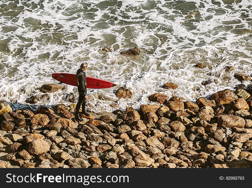 Man Wearing Wetsuit And Holding Red Surfboard On Shore