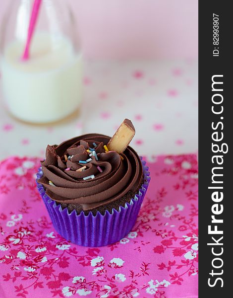 Close up of chocolate cupcake in blue wrapper with sprinkles on pink cloth with bottle of milk.