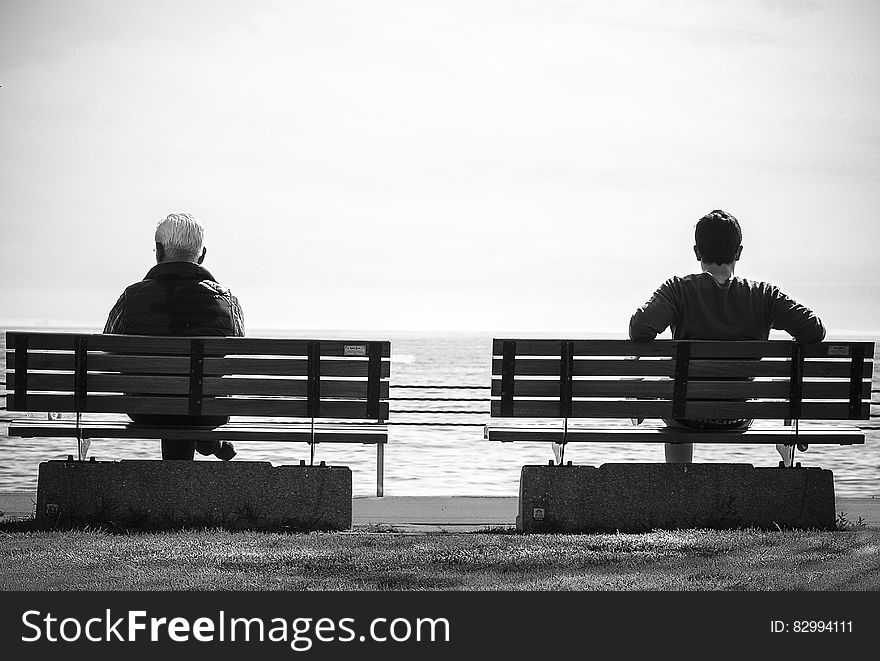 Grayscale Photo 2 Person Sitting in a Separate Benches on the Seaside