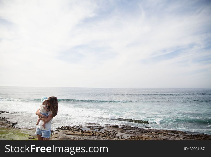 Mother Holding Her Baby in Front of Seashore during Daytime