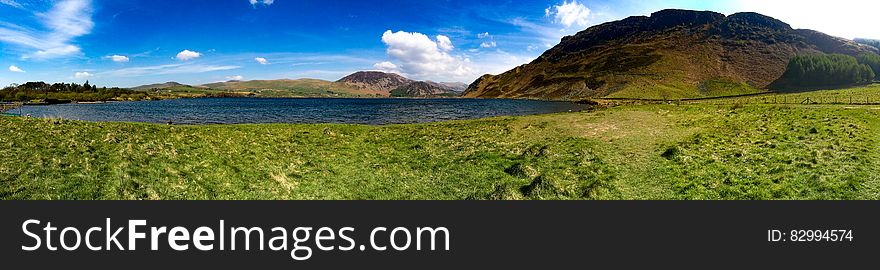 Panoramic view of countryside lake next to hills with blue sky and cloudscape.