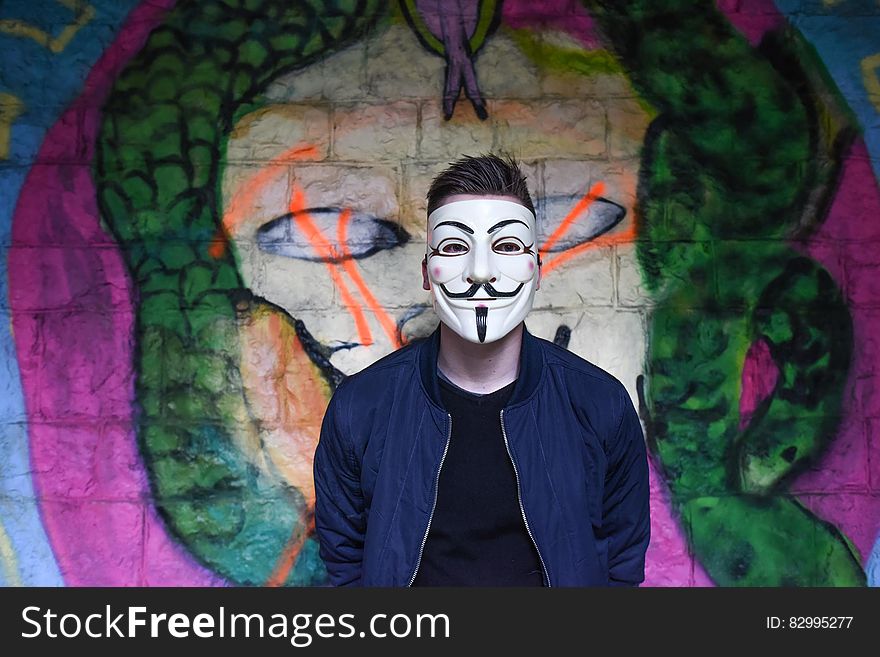 Man in White Mask in Black Crew Neck Shirt and Blue Zip Up Jacket Infront Graffiti Wall