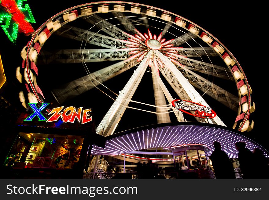 Ferris wheel in funfair at night with motion blur, silhouetted people in foreground.