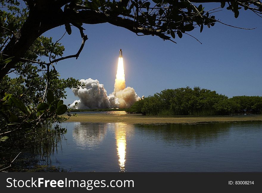 Rocket launch reflecting in calm waters of lake against blue skies.