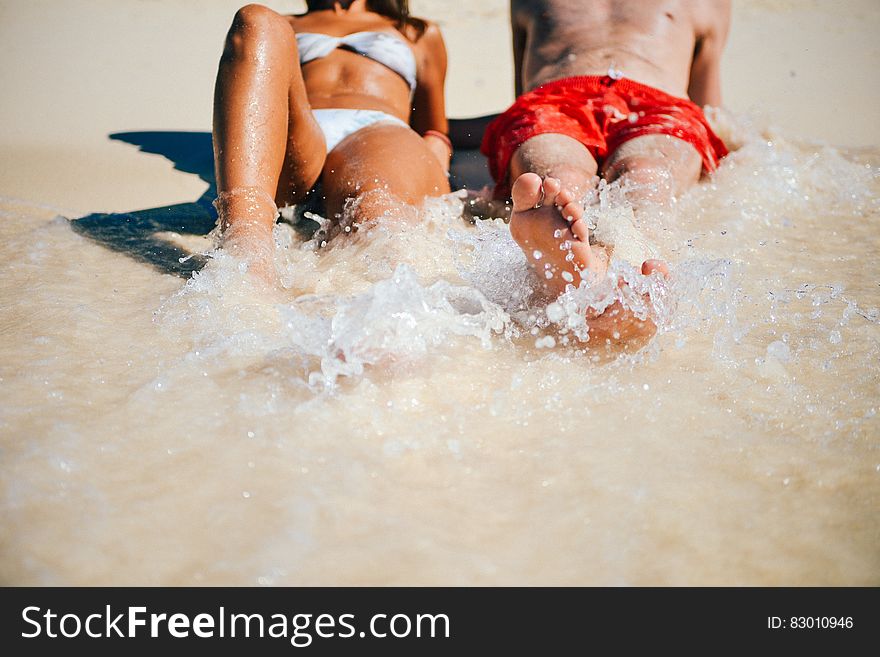 Anonymous couple on the beach both splashing the water with their feet. He is wearing red shorts and she a white bikini looking very sun tanned.