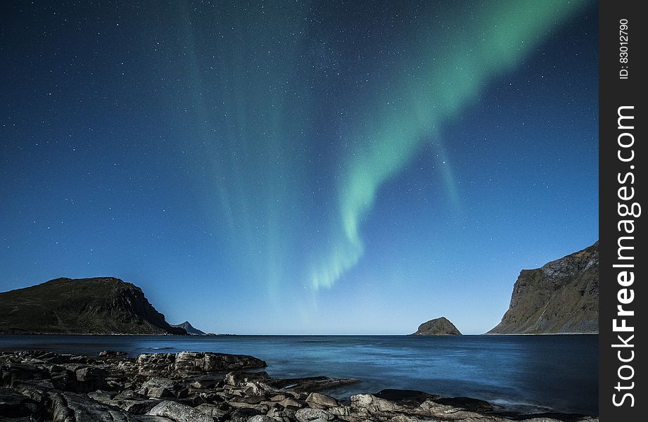 Rocky coastline with islands and cliffs just away from the shore line and the Aurora Borealis a green triangular streak in the blue sky. Rocky coastline with islands and cliffs just away from the shore line and the Aurora Borealis a green triangular streak in the blue sky.