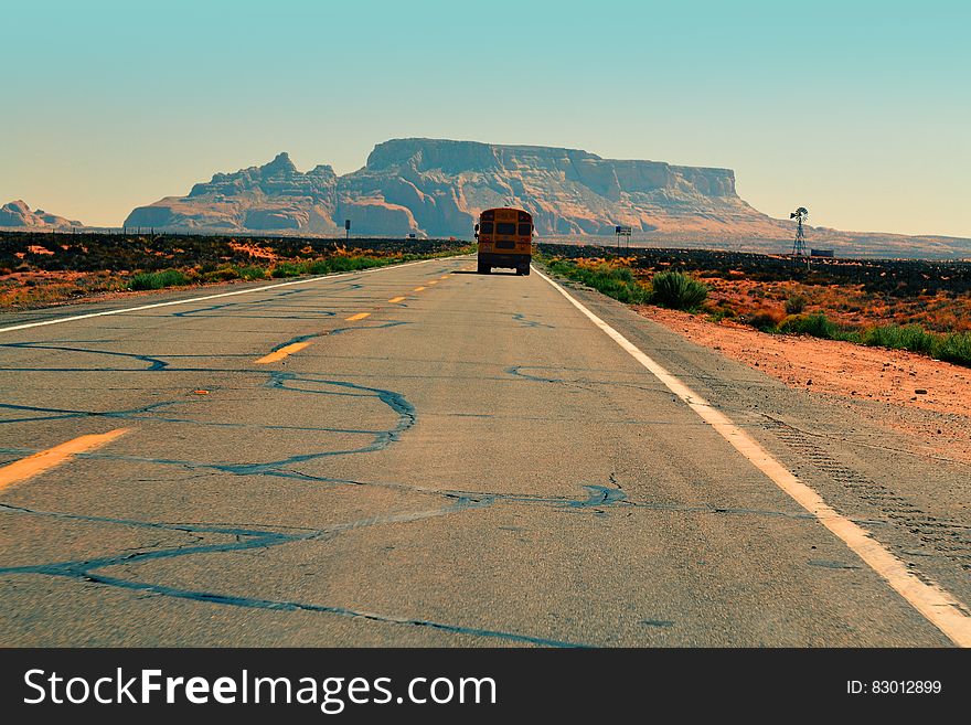 Road through red sandy desert with scrub growing along both sides and yellow bus approaching (or disappearing) . In distance is a table mountain and behind it a pale blue sky. Road through red sandy desert with scrub growing along both sides and yellow bus approaching (or disappearing) . In distance is a table mountain and behind it a pale blue sky.