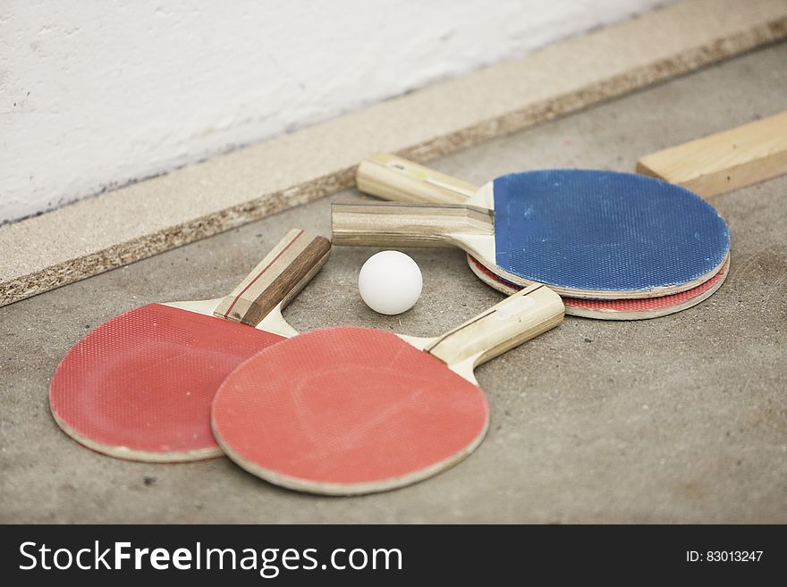 A ping pong ball on the floor with blue and red paddles on the floor. A ping pong ball on the floor with blue and red paddles on the floor.