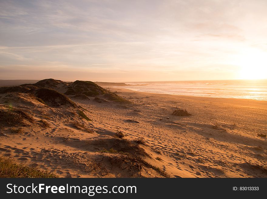 Sand dunes in desert landscape at sunset with sea in background.